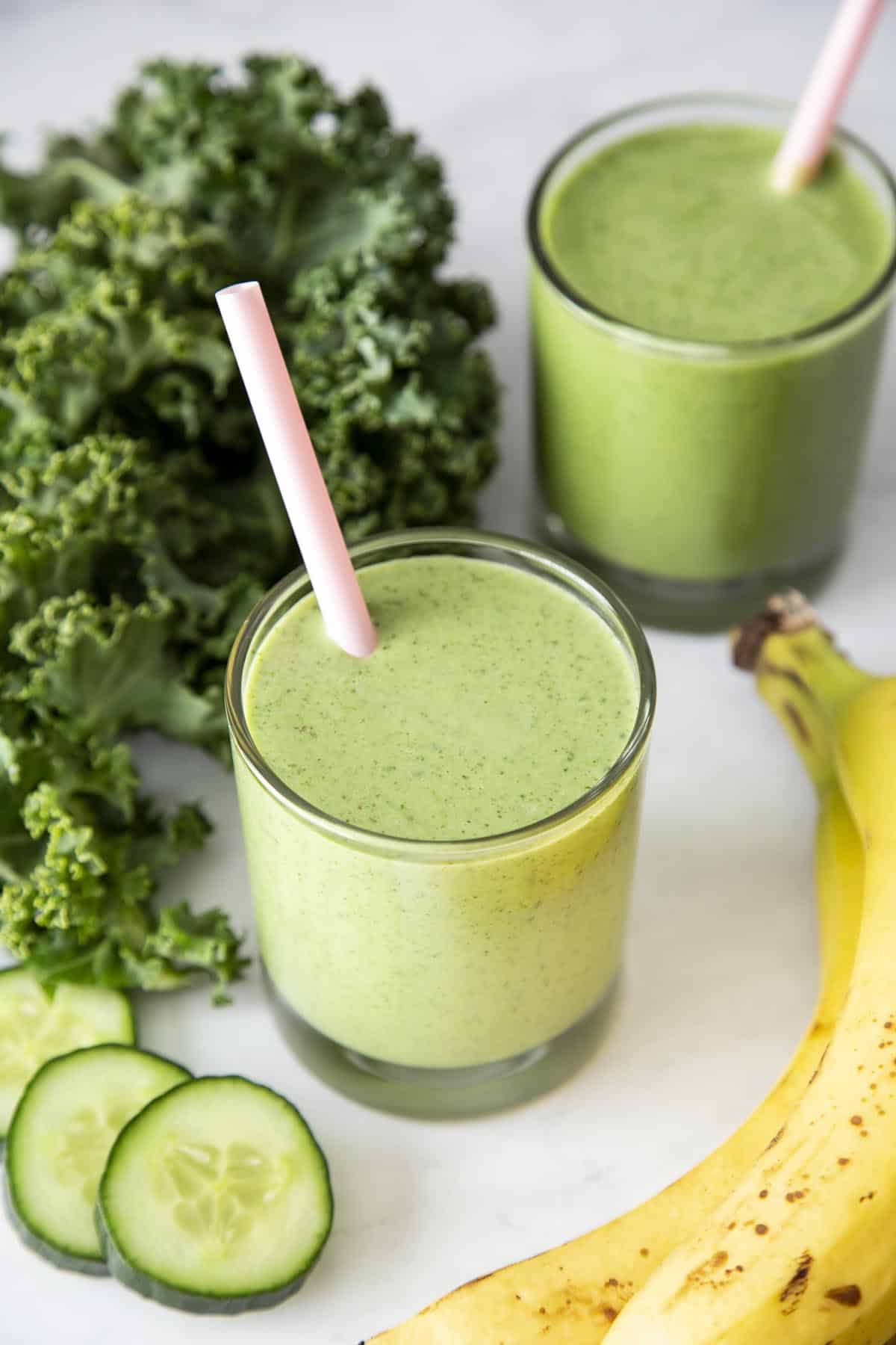 ¾ view of green smoothie in 2 glasses with pink straws next to banana, kale and cucumber slice.