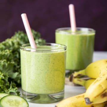 Green smoothie in 2 glasses with pink straws next to banana, kale and cucumber slice.