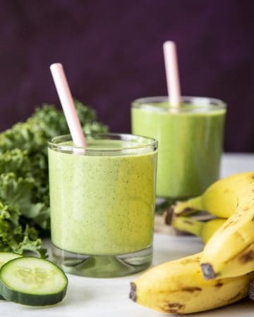 Green smoothie in 2 glasses with pink straws next to banana, kale and cucumber slice.