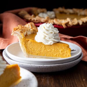 A slice of pumpkin pie on a stack of plates with whipped cream on top.