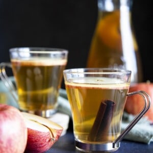 2 glasses of apple ciders next to some apple and a serving jug.