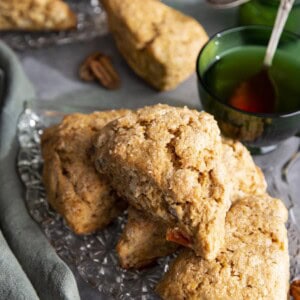 A pile of scones on a glass plate with a green tea cup, more scones in the background.