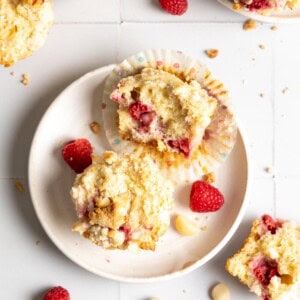 A full raspberry white chocolate muffin and a half on a white plate with a couple raspberries.