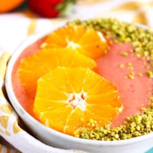 Pink strawberry orange juice smoothie bowl garnished with orange slices and pistachio.