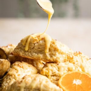 A spoon drizzling glaze over a clementine coconut scone sitting on top of a plate of scones.