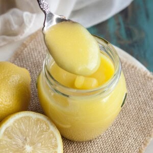 A spoon picking up some lemon curd from a jar next to some lemons