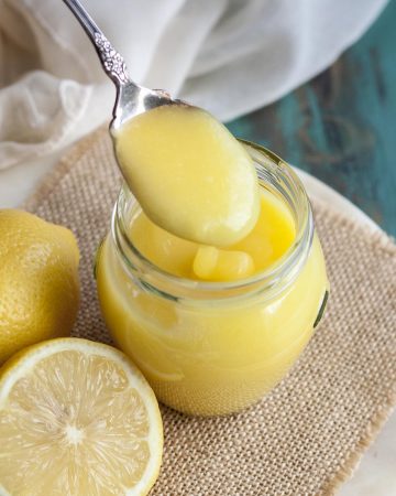 A spoon picking up some lemon curd from a jar next to some lemons