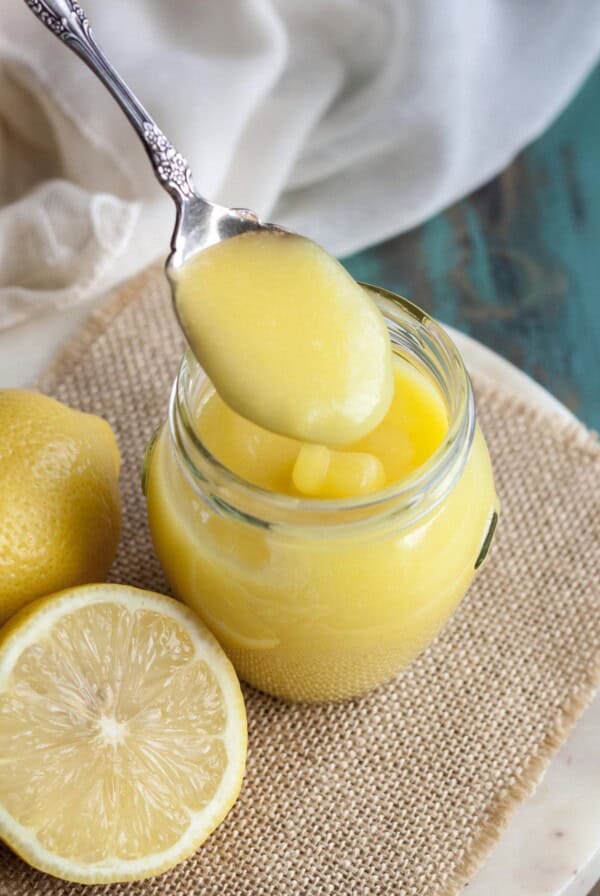A spoon picking up some lemon curd from a jar next to some lemons