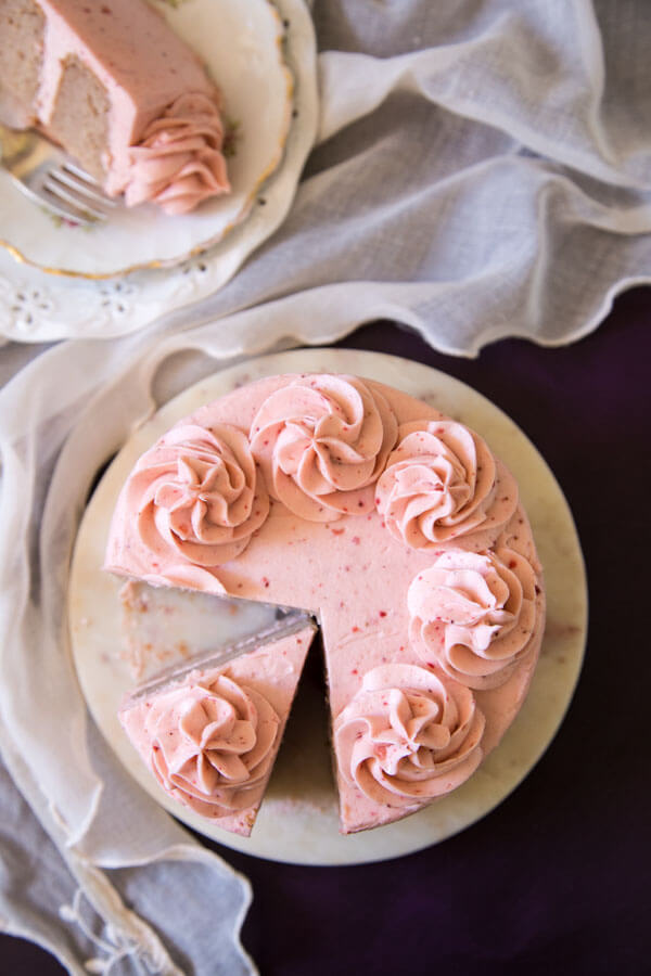 Strawberry cake with strawberry cream cheese frosting on a cake stand next to a plate with a slice of cake