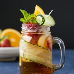 A glass of Pimm's cup cocktail garnished with cucumber, strawberry and orange slice on a blue table top.