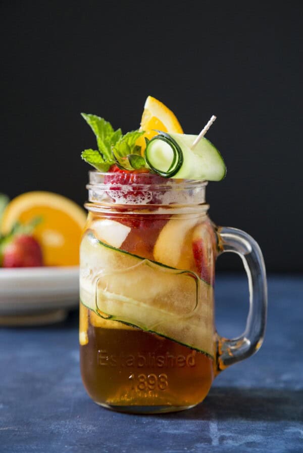 A glass of Pimm's cup cocktail garnished with cucumber, strawberry and orange slice on a blue table top.