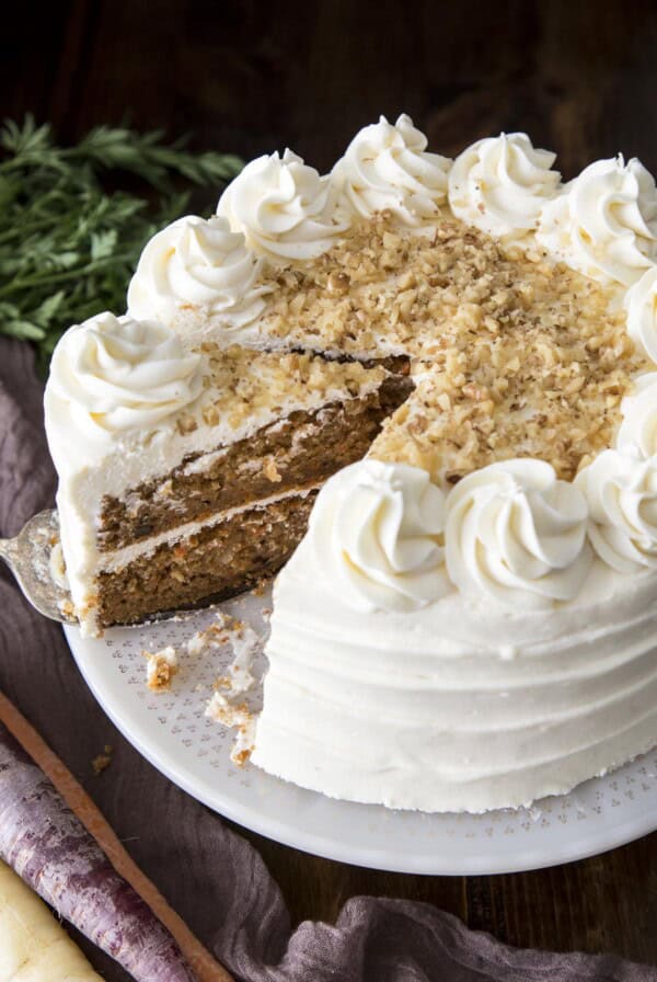 A carrot cake on a cake stand with a wedge taken out.