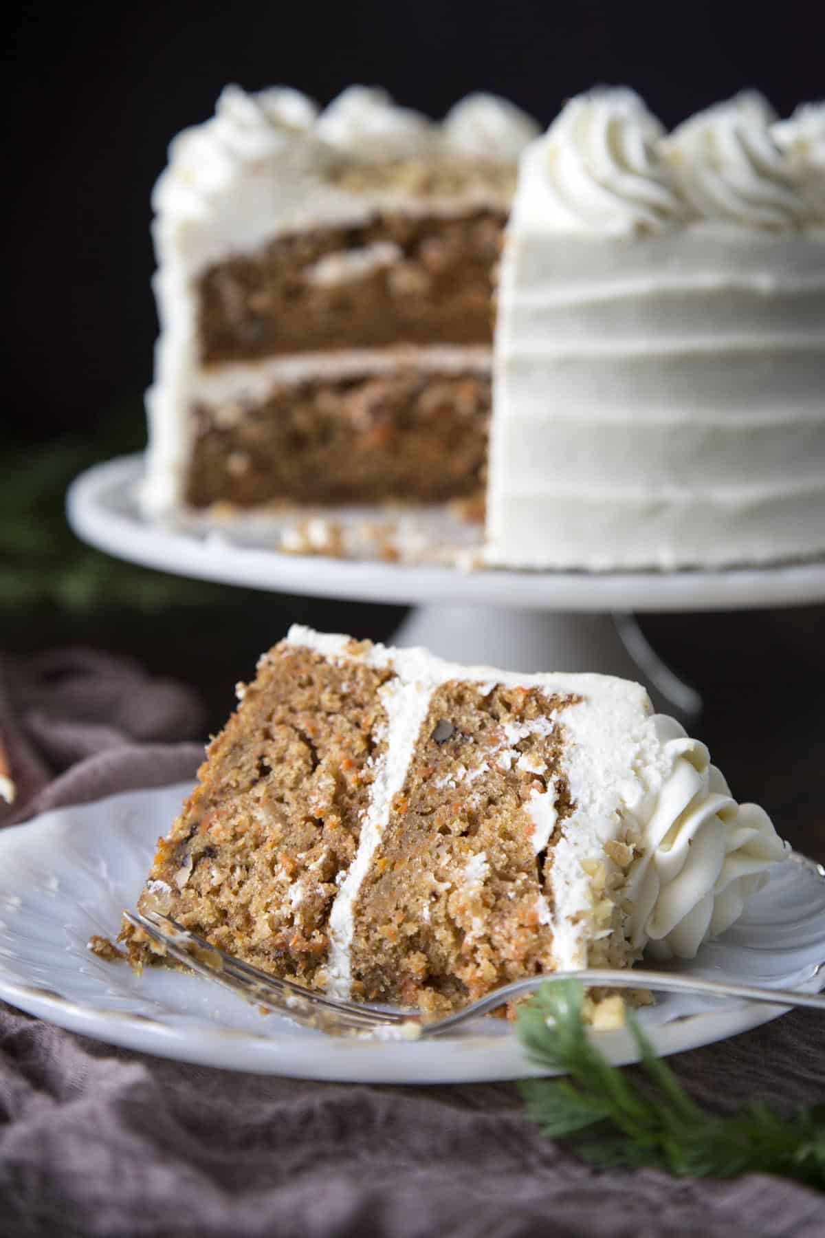 A slice of carrot cake on a plate with the rest of the cake in the back.