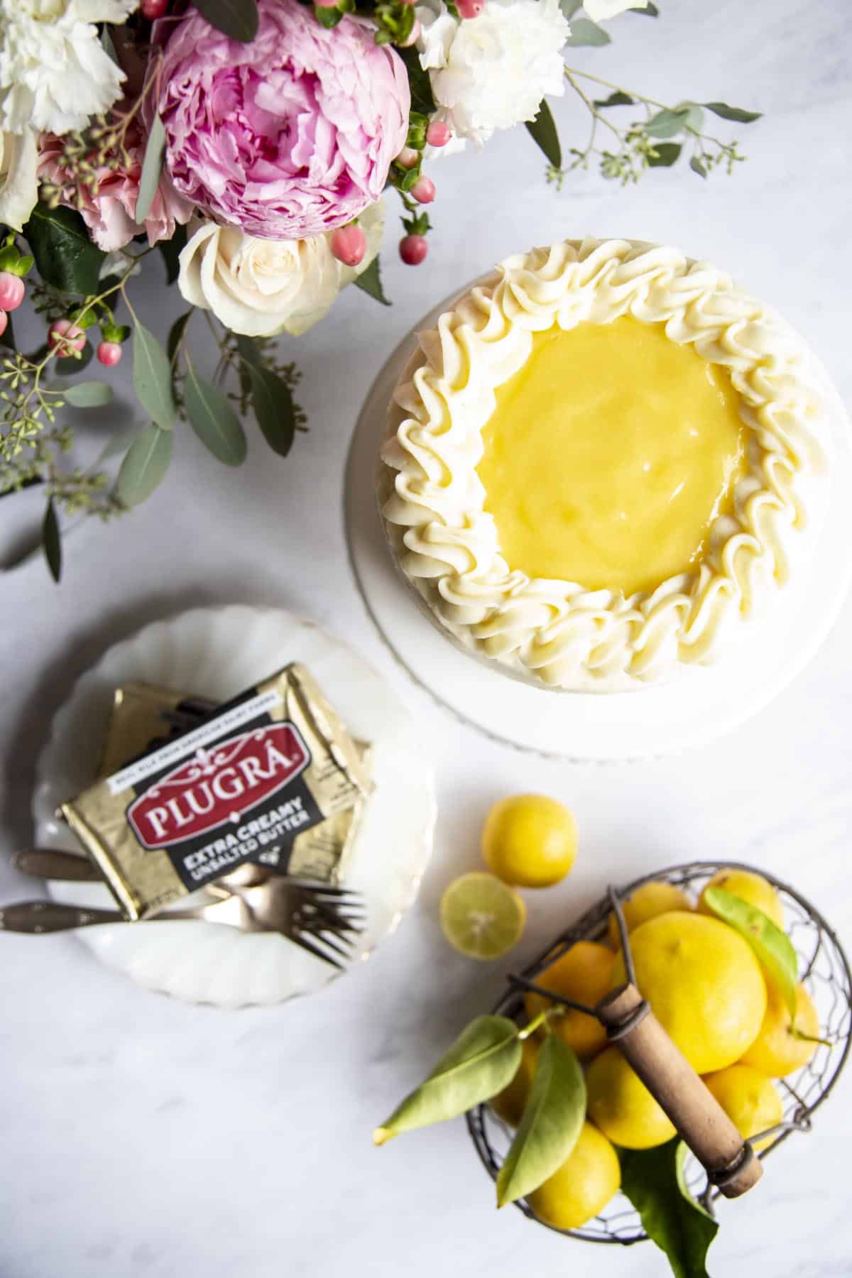 Lemon cake on a cake stand next to some flowers, a basket of lemon and a block of Plugra butter