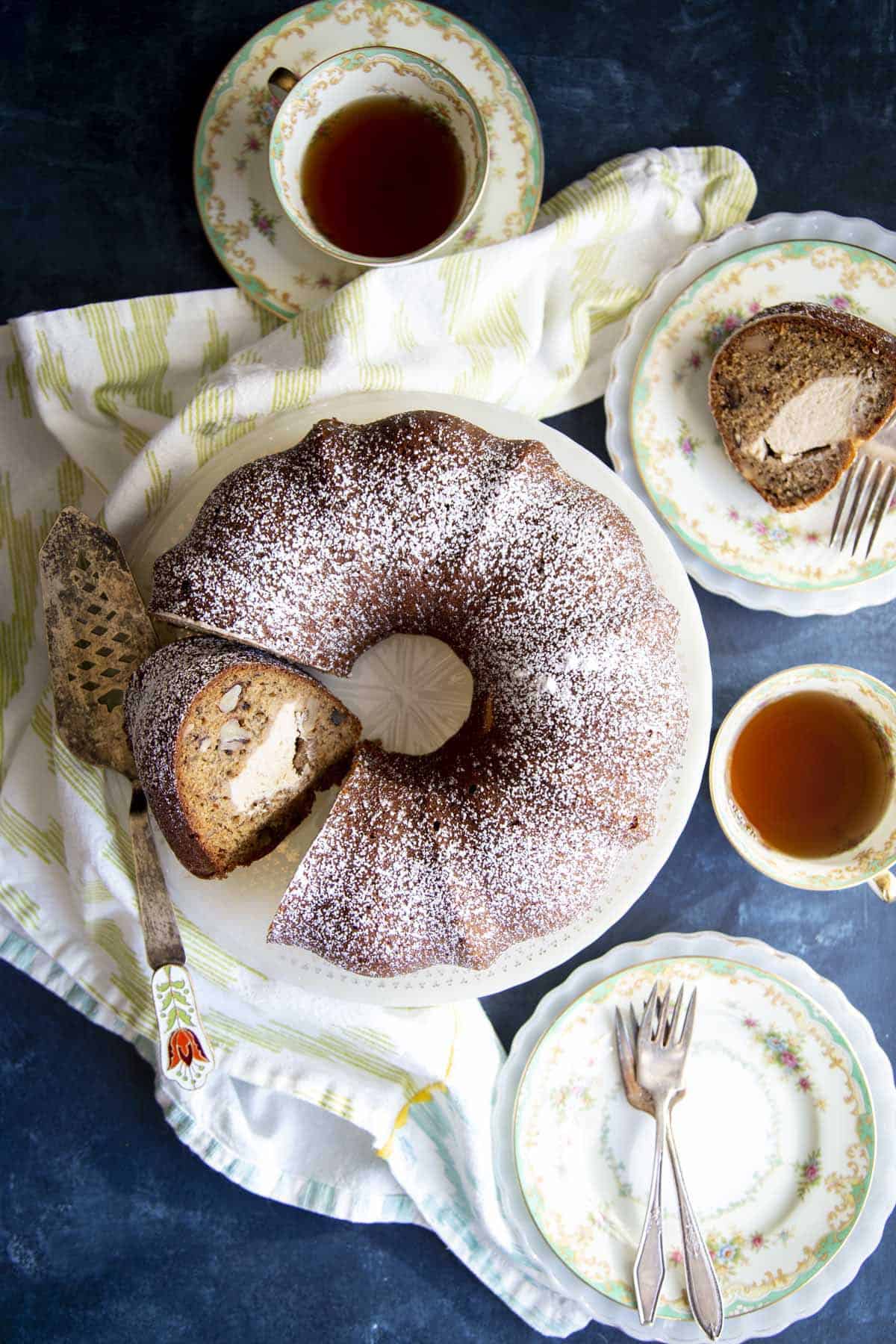 A sliced banana bundt cake on a cake stand, with tea and cake slice on plates surrounding it.