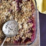 Apple and plum crumble in a glass dish with a serving spoon in it.