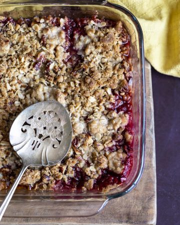 Apple and plum crumble in a glass dish with a serving spoon in it.