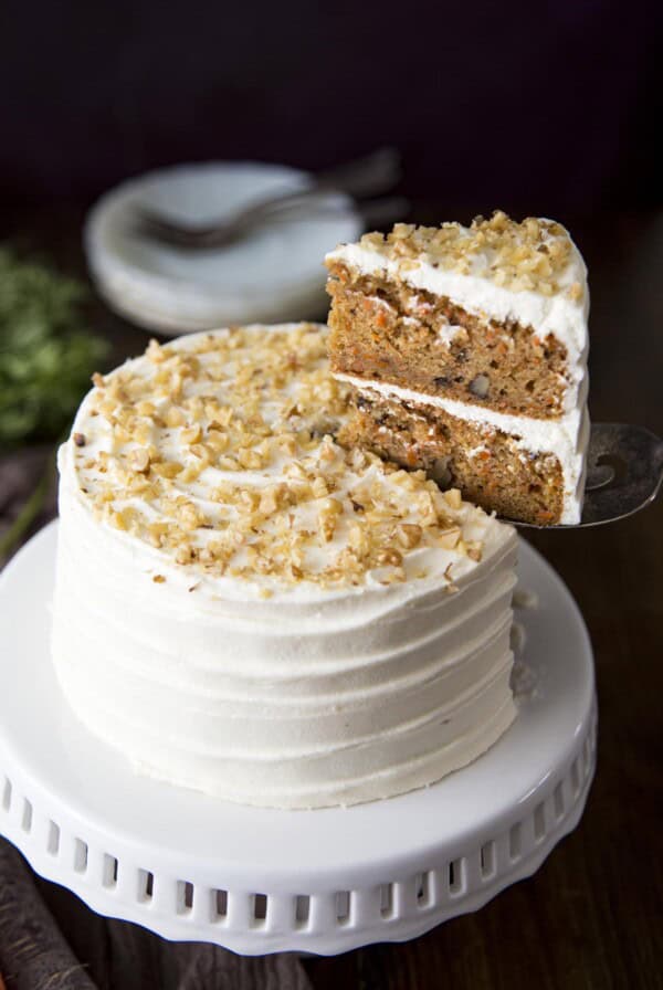 A mini carrot cake on a white cake stand with a slice being cut.