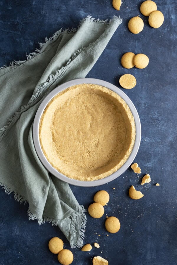 A vanilla wafer crust on a blue table top next to a green towel and vanilla wafer cookies all around.