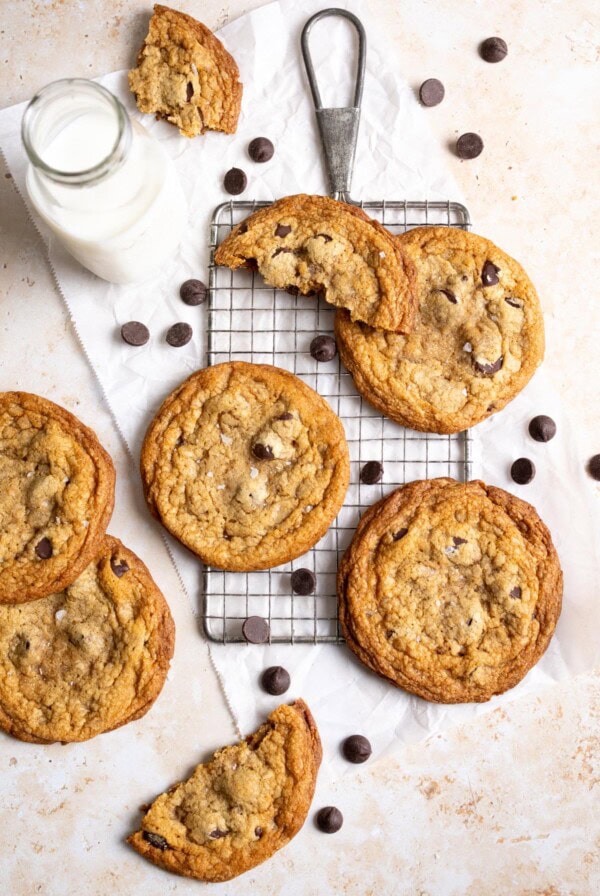Toffee chocolate chip cookies spread on a table top with chocolate chips scattered around next to a bottle of milk.