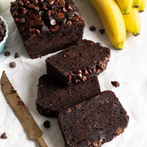 A loaf of chocolate chocolate chip banana bread, sliced, next to some bananas.