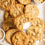 A pile of toffee macadamia oatmeal cookies, in the middle is a glass of milk with a half eaten cookie resting on top.