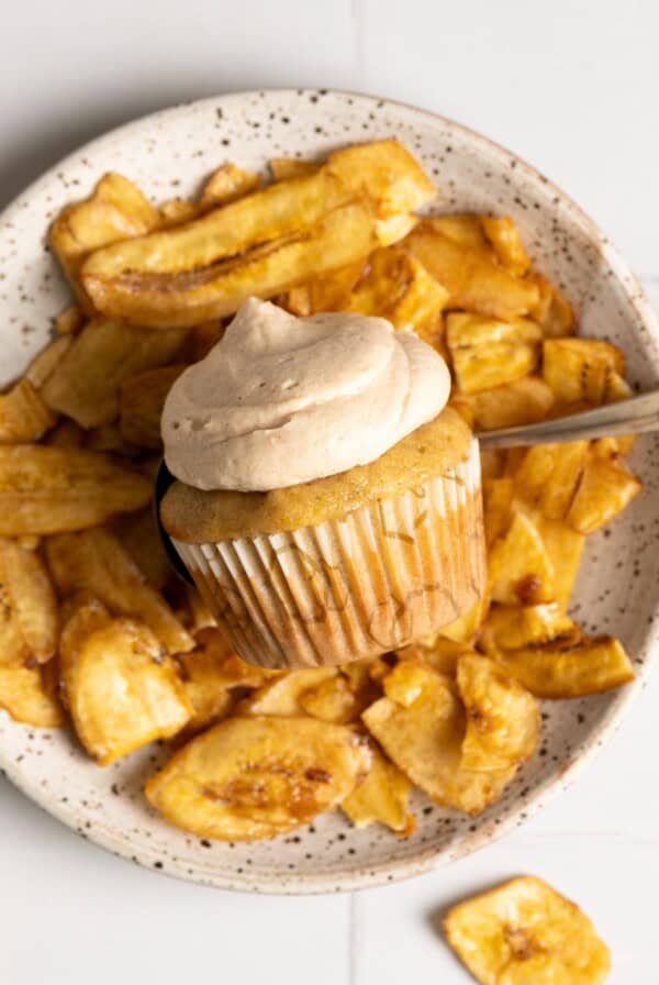 A miso banana cupcake with cinnamon cream cheese frosting laying on top of a plate of banana chips.