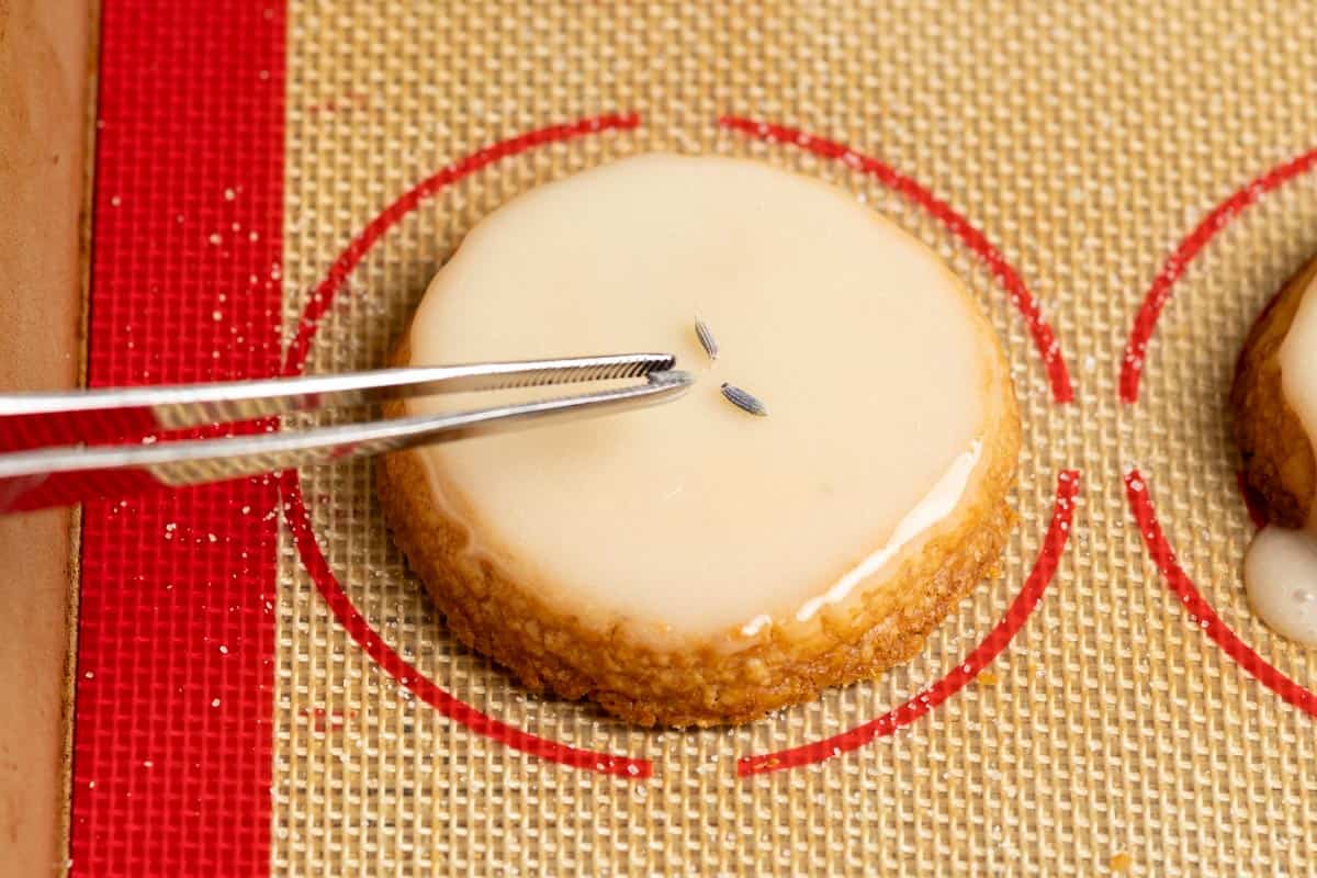 Placing a dried lavender flower on top of a glazed cookie with a tweezer.