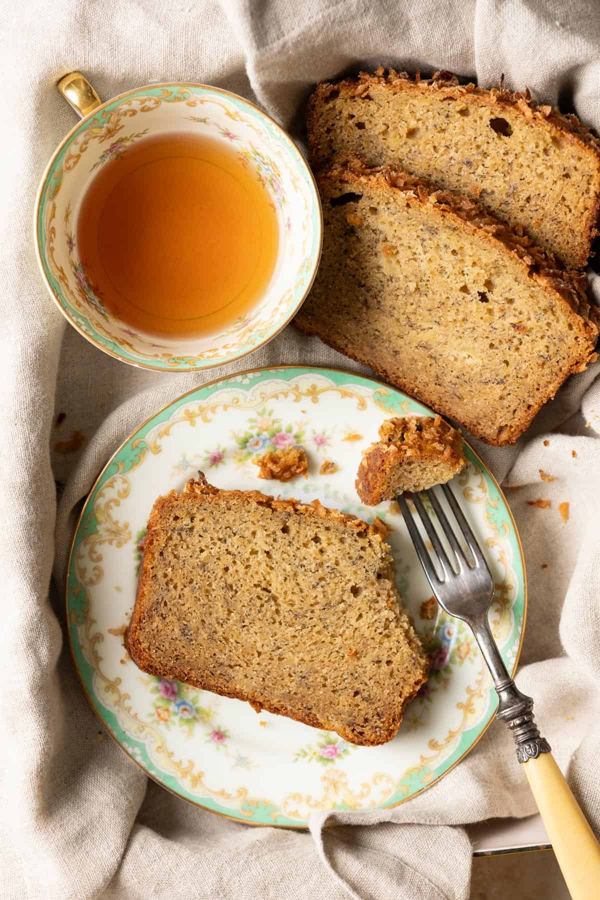 A slice of pineapple banana bread on a plate with a fork, next to a tea cup and 2 more slices.